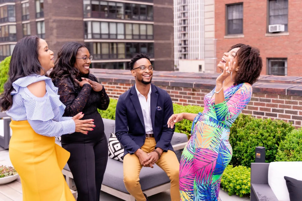 Four people are socializing and laughing on a rooftop patio with city buildings in the background.