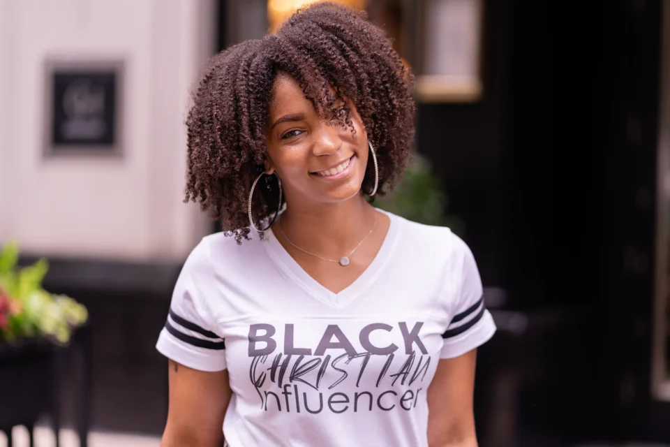 A woman with curly hair smiles at the camera, wearing hoop earrings and a white T-shirt that reads "Black Christian Influencer.
