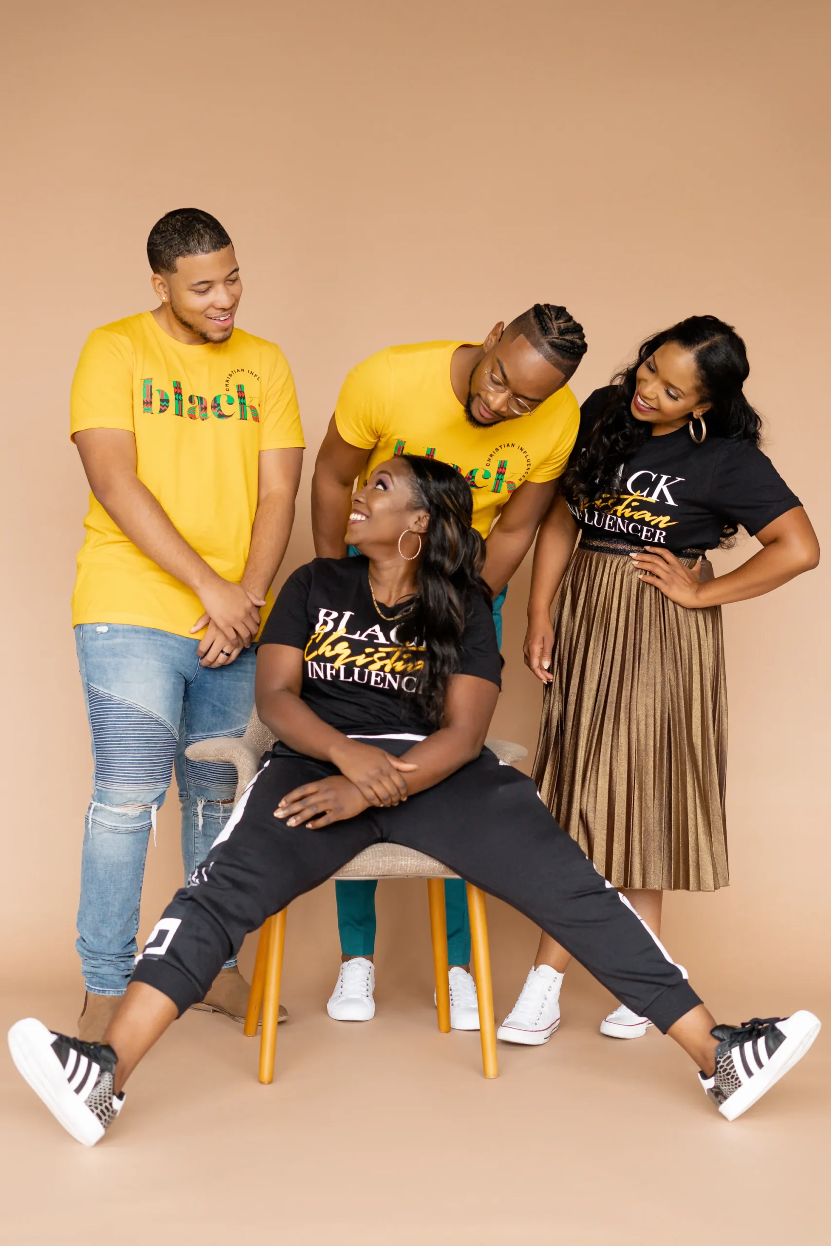Four people pose against a tan background; three stand smiling behind a seated woman with legs extended. They wear coordinated yellow and black outfits with graphic text.