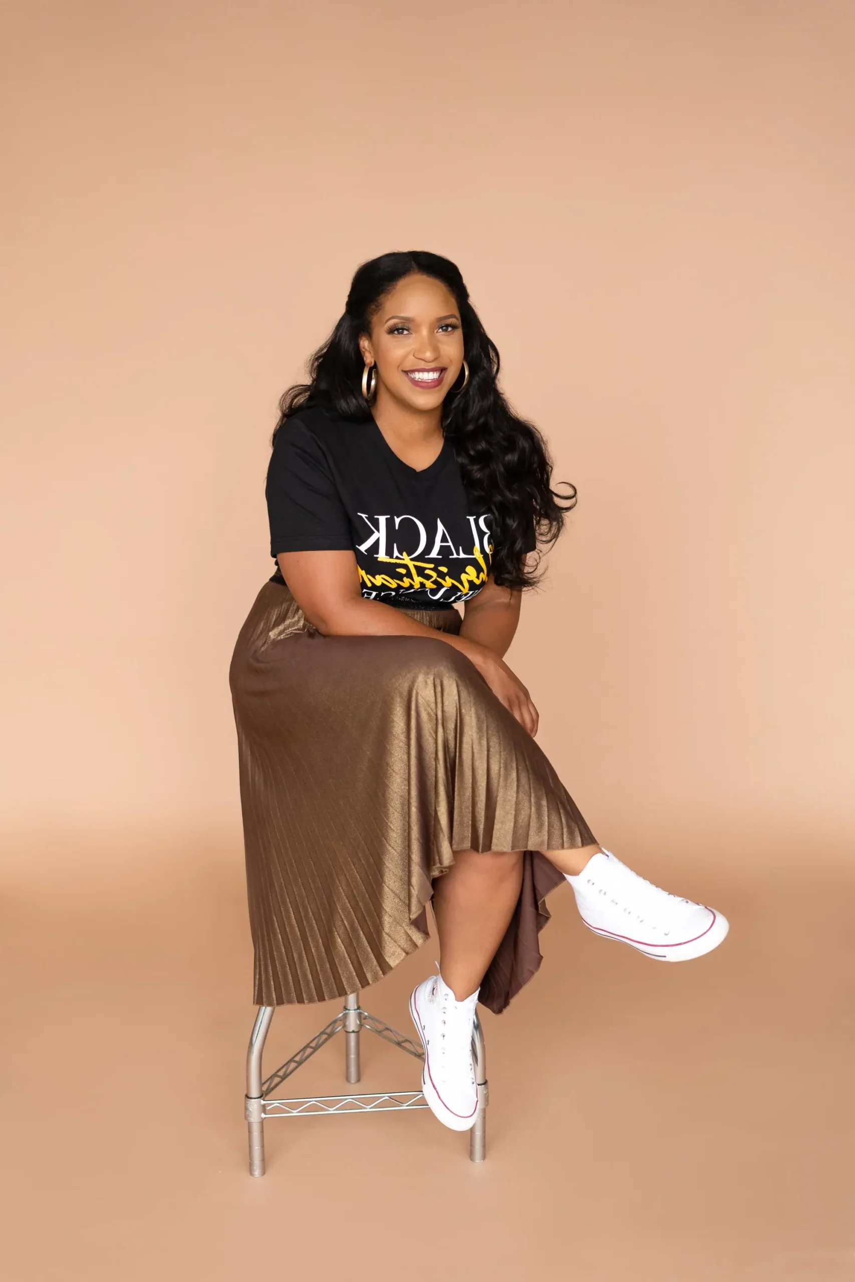 Woman with long dark hair sits on a stool against a beige background, wearing a "Black Christian" t-shirt, metallic pleated skirt, and white sneakers, smiling at the camera.
