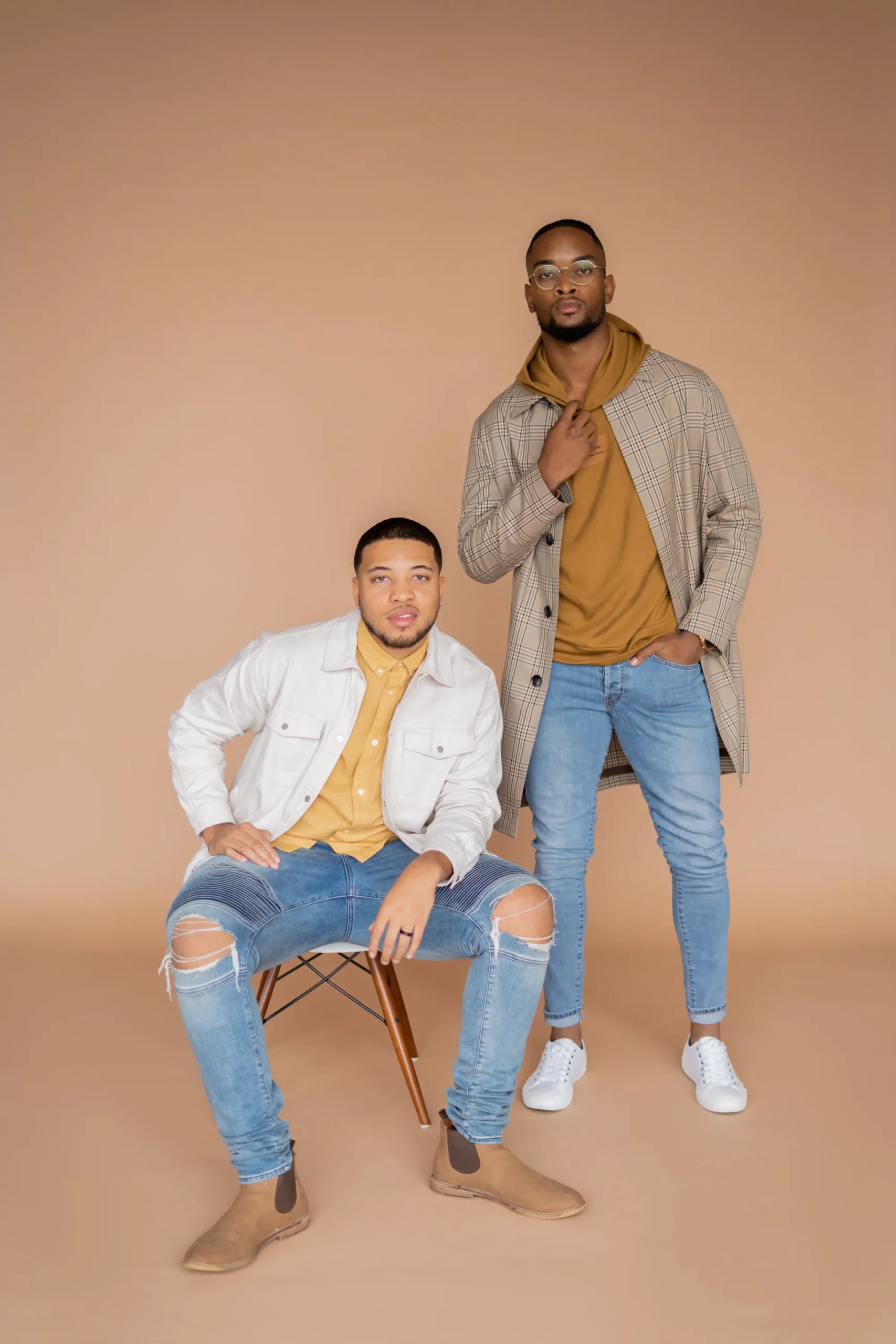 Two men posing against a beige backdrop; one is seated wearing a white jacket and ripped jeans, the other is standing wearing a plaid coat, mustard hoodie, and jeans.