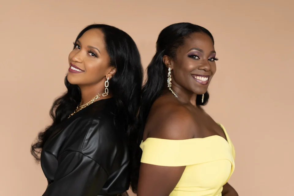 Two women with long dark hair stand back to back, smiling at the camera. One wears a black outfit, the other a yellow off-the-shoulder dress, both with statement earrings.