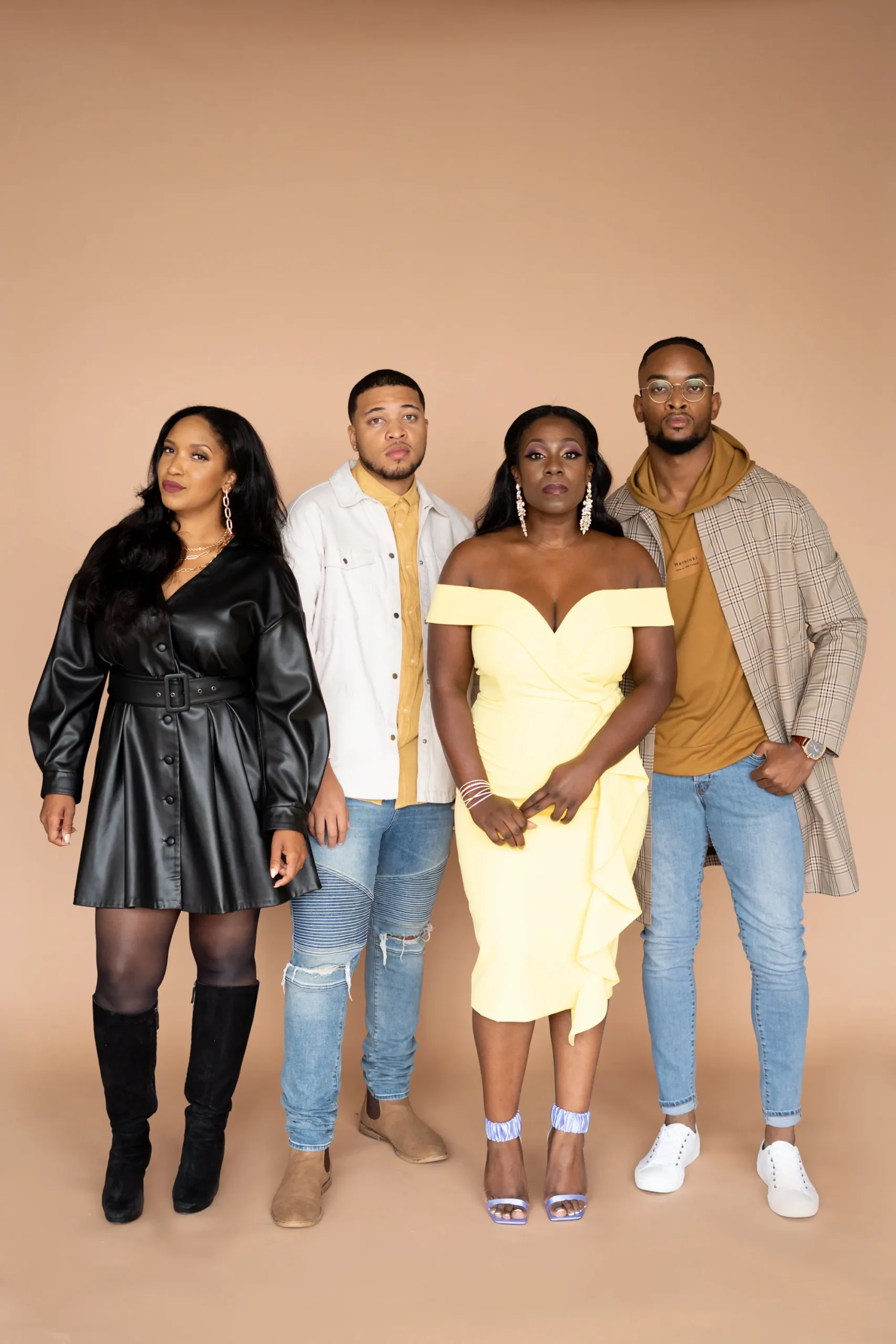 Four people stand together against a tan backdrop, dressed in stylish semi-formal outfits, looking directly at the camera.