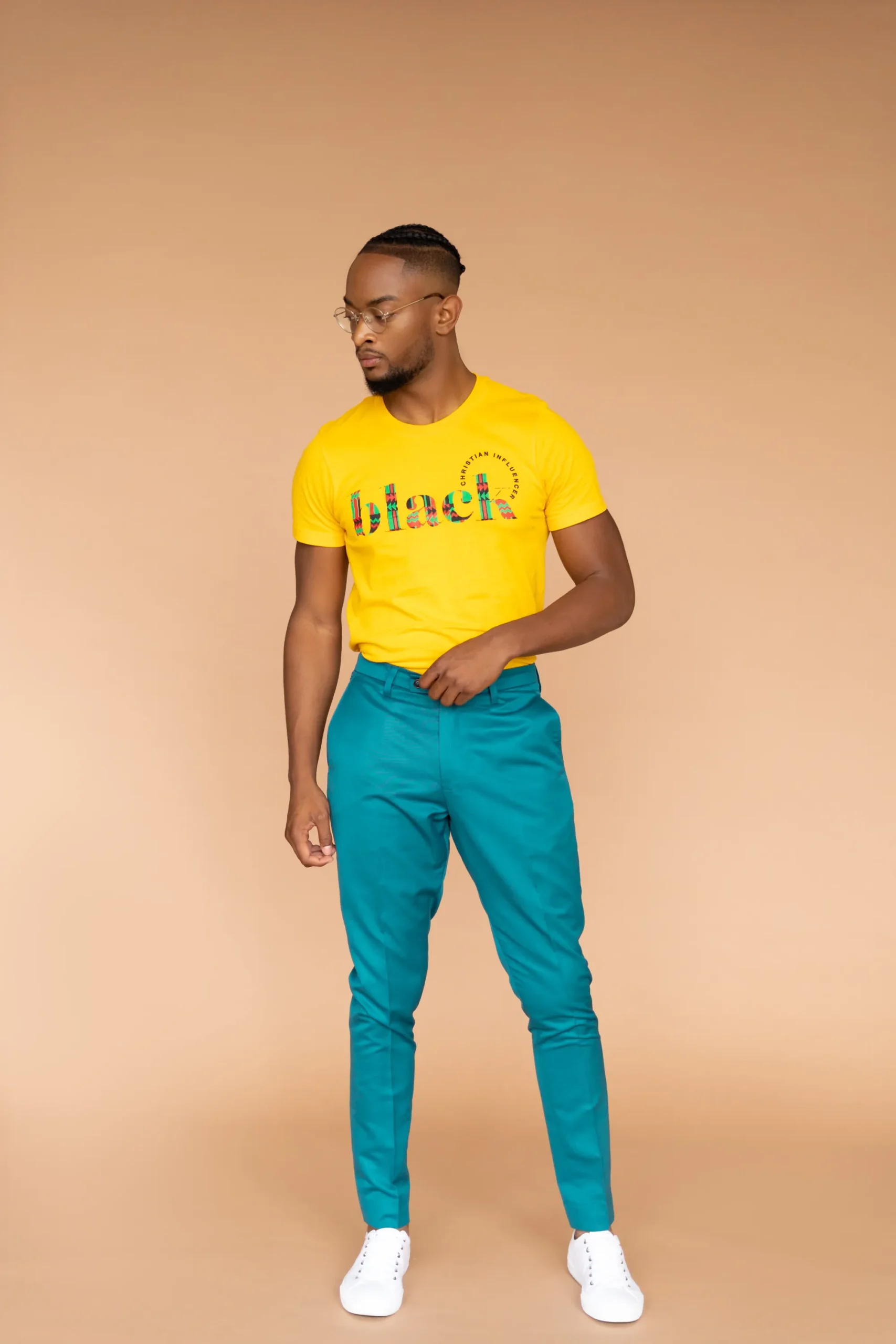 A man wearing a yellow t-shirt, teal pants, and white sneakers stands against a plain beige background.