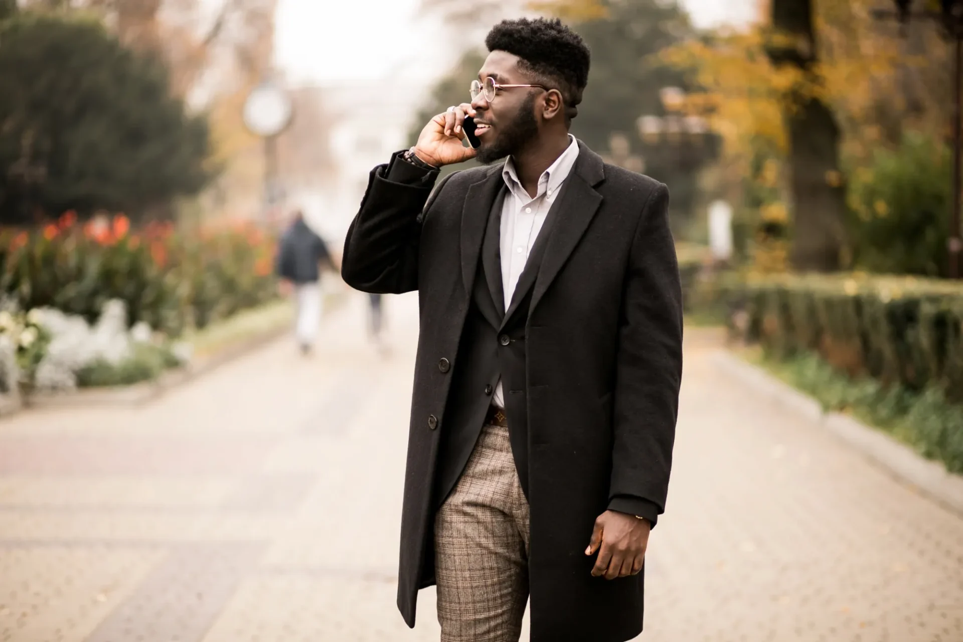 A man wearing a dark coat and plaid pants is walking outdoors on a paved path, talking on his phone. Trees and blurred pedestrians are visible in the background.