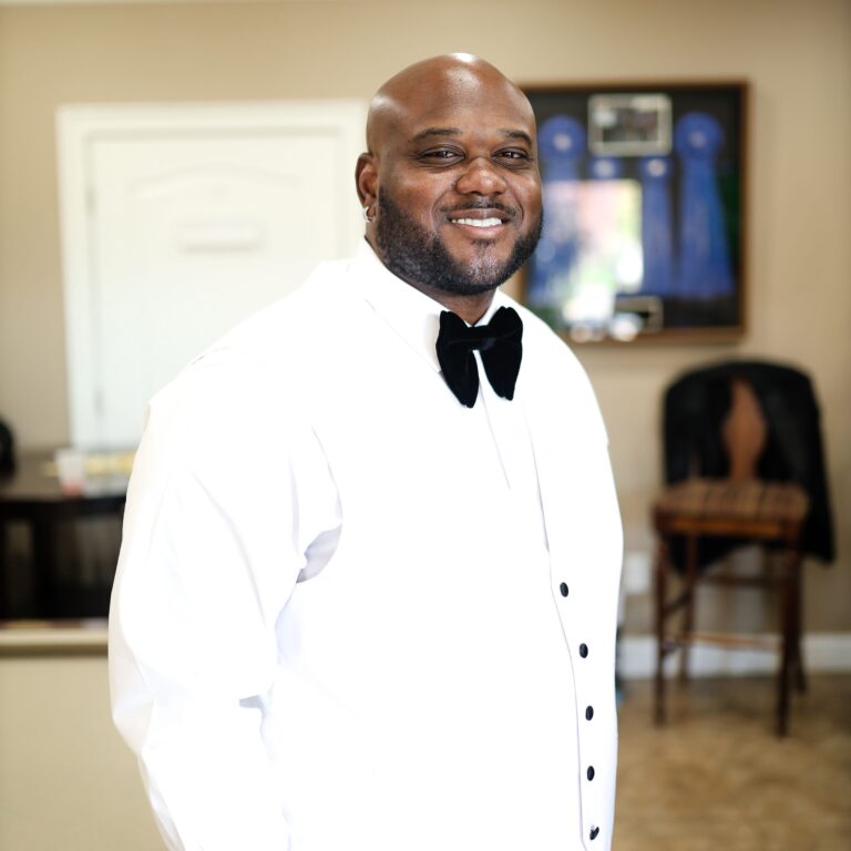 A man wearing a white shirt, black bow tie, and black buttons stands indoors, smiling at the camera. The background includes a chair, a coat, and framed items on the wall.