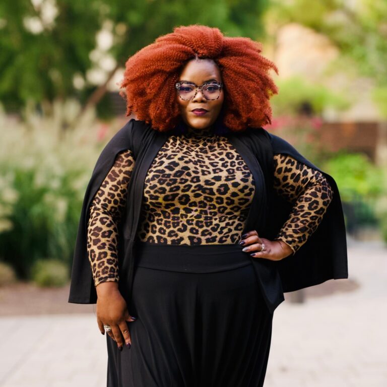 A person with voluminous red hair, wearing glasses, a leopard print top, a black cape, and black skirt stands confidently outdoors on a paved path.