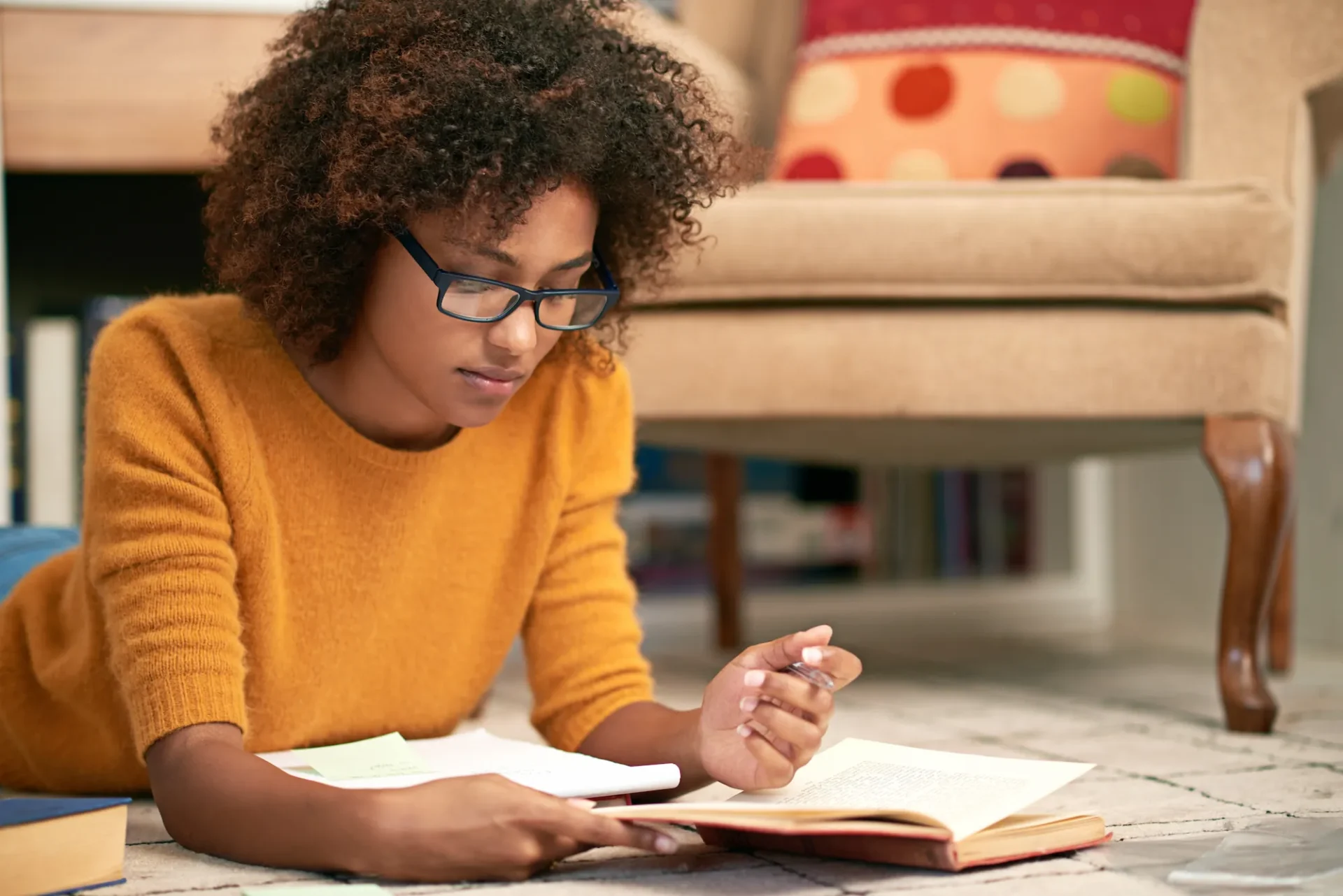 A woman wearing glasses and a yellow sweater lies on the floor reading a book, with papers and a pen in hand, near a beige chair.